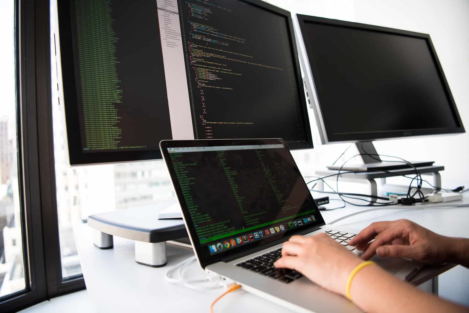 A imagem, utilizada para ilustrar como é possível ser um programador melhor, conta com as mãos de uma pessoa desenvolvendo um software em um computador sobre uma mesa com três telas alternativas